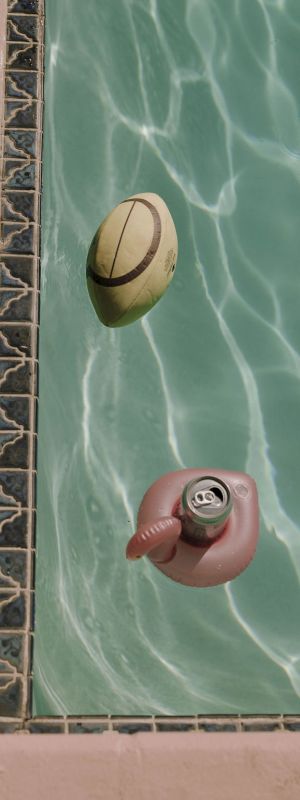 A football and a can in a flamingo float are floating in a swimming pool with a tiled edge, under clear sunlight.