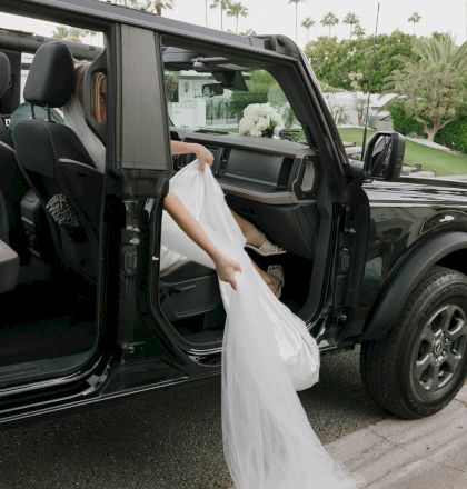 A person in a wedding dress exits a vehicle, holding the dress. Palm trees are in the background.