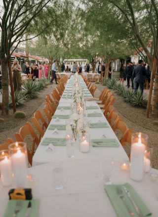 A long outdoor dining table is elegantly set with candles and place settings. People mingle in the background among trees and plants.