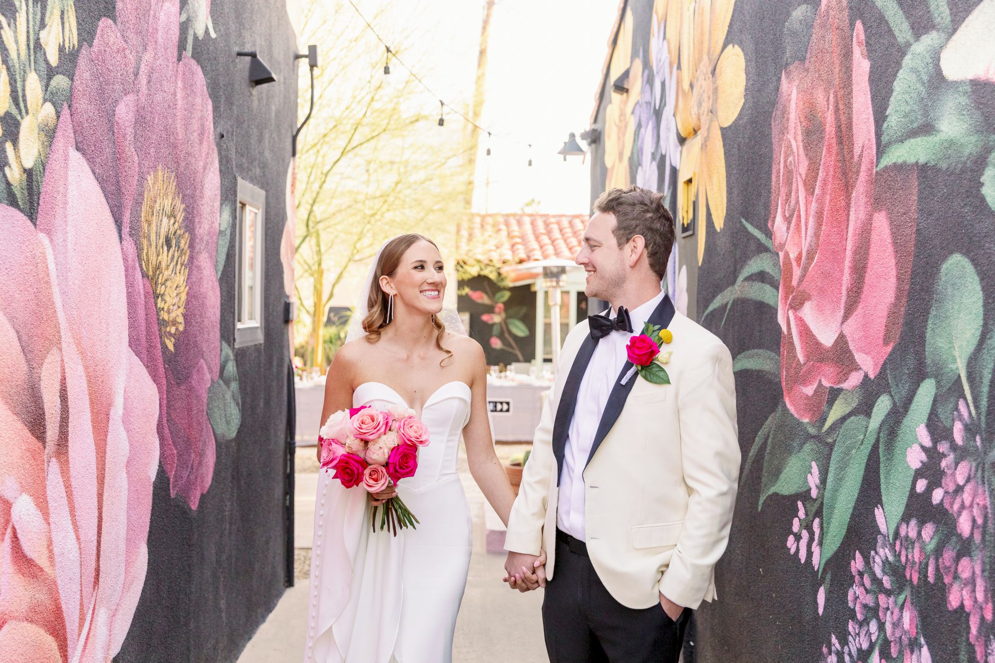 A couple in wedding attire walks hand-in-hand in a floral-painted alley, smiling at each other.