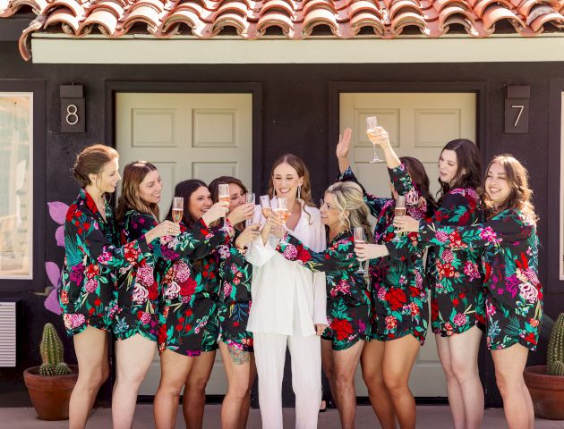 A group of women in floral robes celebrating, holding glasses, and standing outside in front of numbered doors.