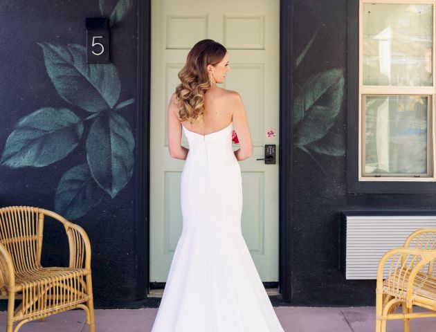 A woman in a white dress stands in front of a door with a number 5, next to a wicker chair and mural leaves.