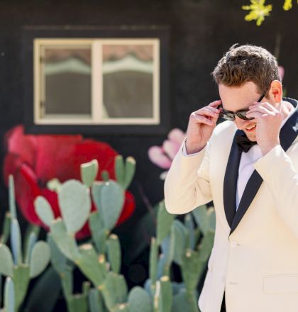 A man in a tuxedo adjusts his sunglasses, standing next to a mural featuring red flowers and cacti.