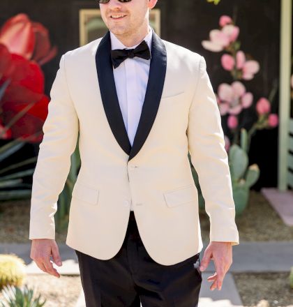 A person in a cream tuxedo jacket with black lapels, bow tie, and sunglasses, standing outdoors near vibrant floral decorations.