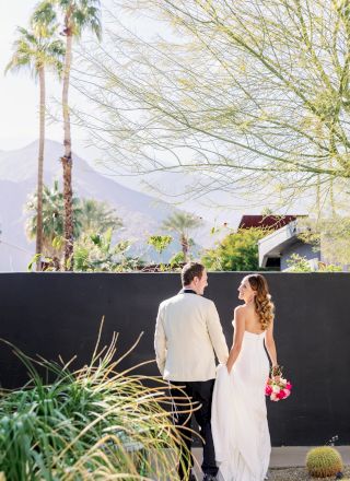 A couple in wedding attire strolls hand in hand, surrounded by desert plants and mountains, beneath a clear sky.