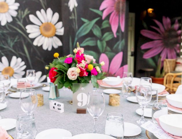 A beautifully set outdoor table with gold cutlery, pink napkins, and colorful flowers against a mural of daisies and large pink flowers.