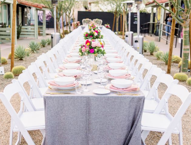 An elegantly set outdoor banquet table with white chairs, gray tablecloths, and floral centerpieces, surrounded by desert landscaping.