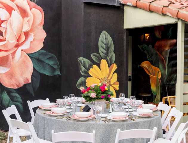 A round table is set with white chairs and tableware, surrounded by a wall with colorful floral murals.