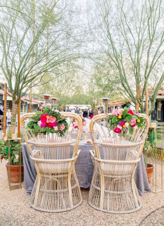 Outdoor dining setup with rattan chairs adorned with colorful flowers, a grey tablecloth, and trees in the background.