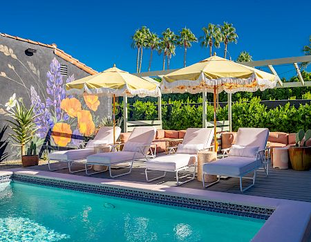 A poolside area with lounge chairs, umbrellas, potted plants, and a mural on the wall. Palm trees are visible in the background.