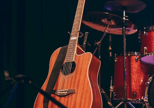 An acoustic guitar stands on stage with a drum set in the background, ready for a musical performance.