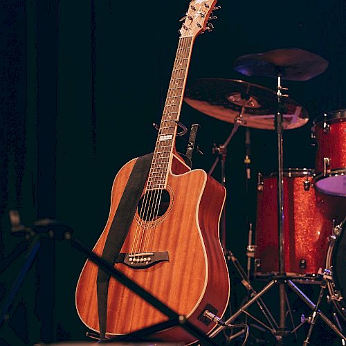 An acoustic guitar stands on stage with a drum set in the background, ready for a musical performance.