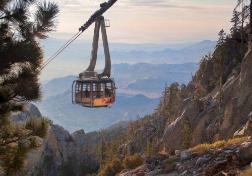 An aerial tramway travels through a mountainous landscape with a vast view of valleys and distant peaks, surrounded by trees and rocks.