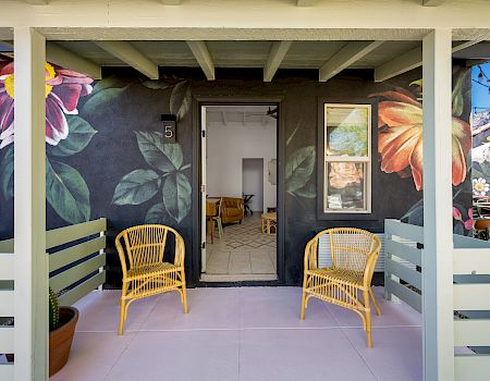 A porch with two yellow chairs, floral murals on walls, and an open door leading inside.