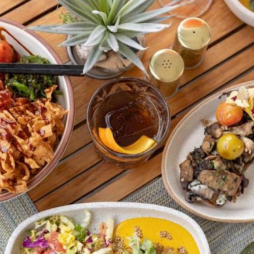 A variety of colorful dishes, including salads and meat, with a drink and garnished with flowers on a wooden table setting.