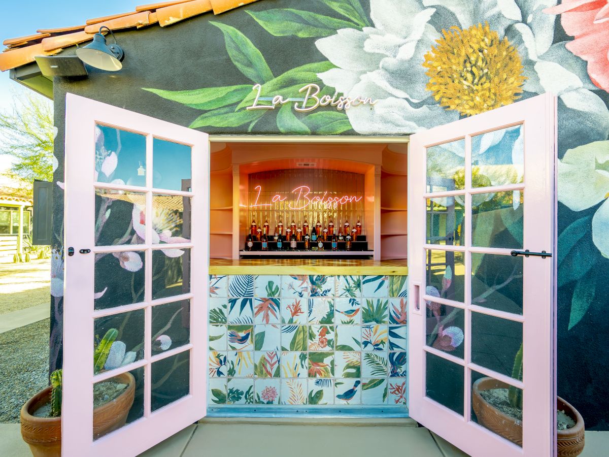 The image shows a floral-themed bar with pink doors open, revealing bottles on display under neon lights reading "La Botica."