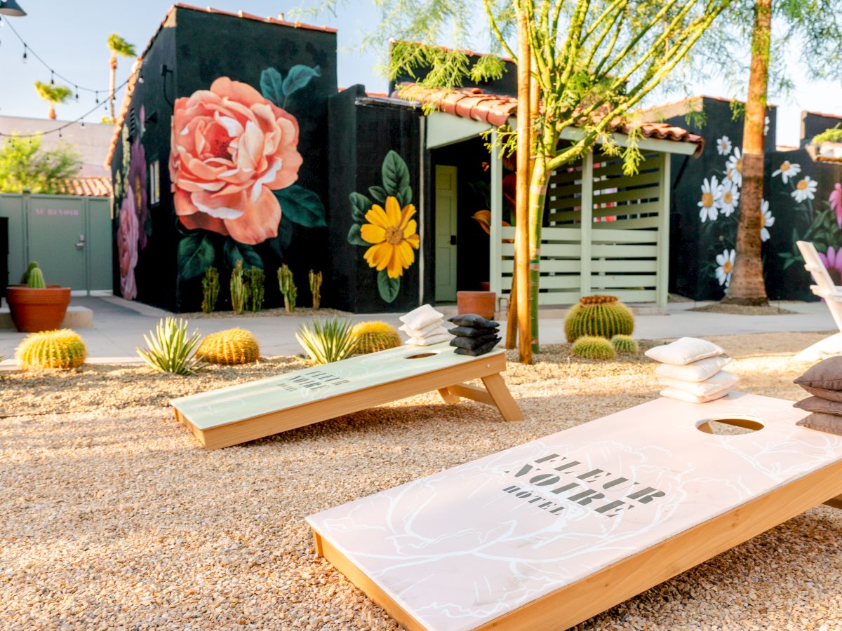 Outdoor scene with cornhole boards, painted murals of flowers, desert plants, and string lights on a sunny day.