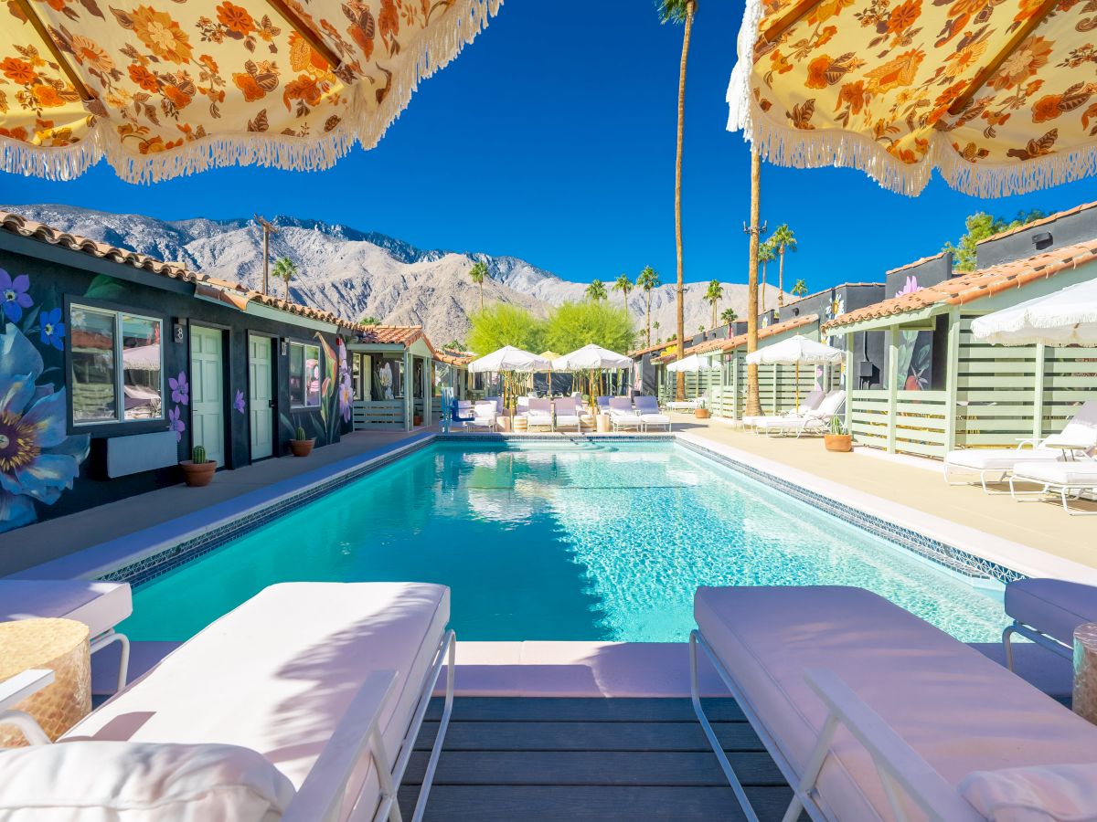 A sunny poolside scene with lounge chairs, floral parasols, and mountains in the background.