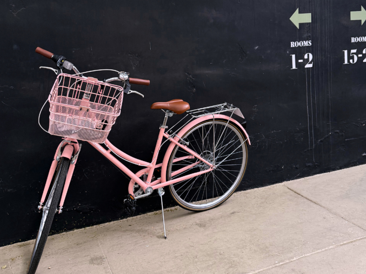 A pink bicycle with a basket is against a black wall with arrows pointing to rooms 1-2 and 15-21.
