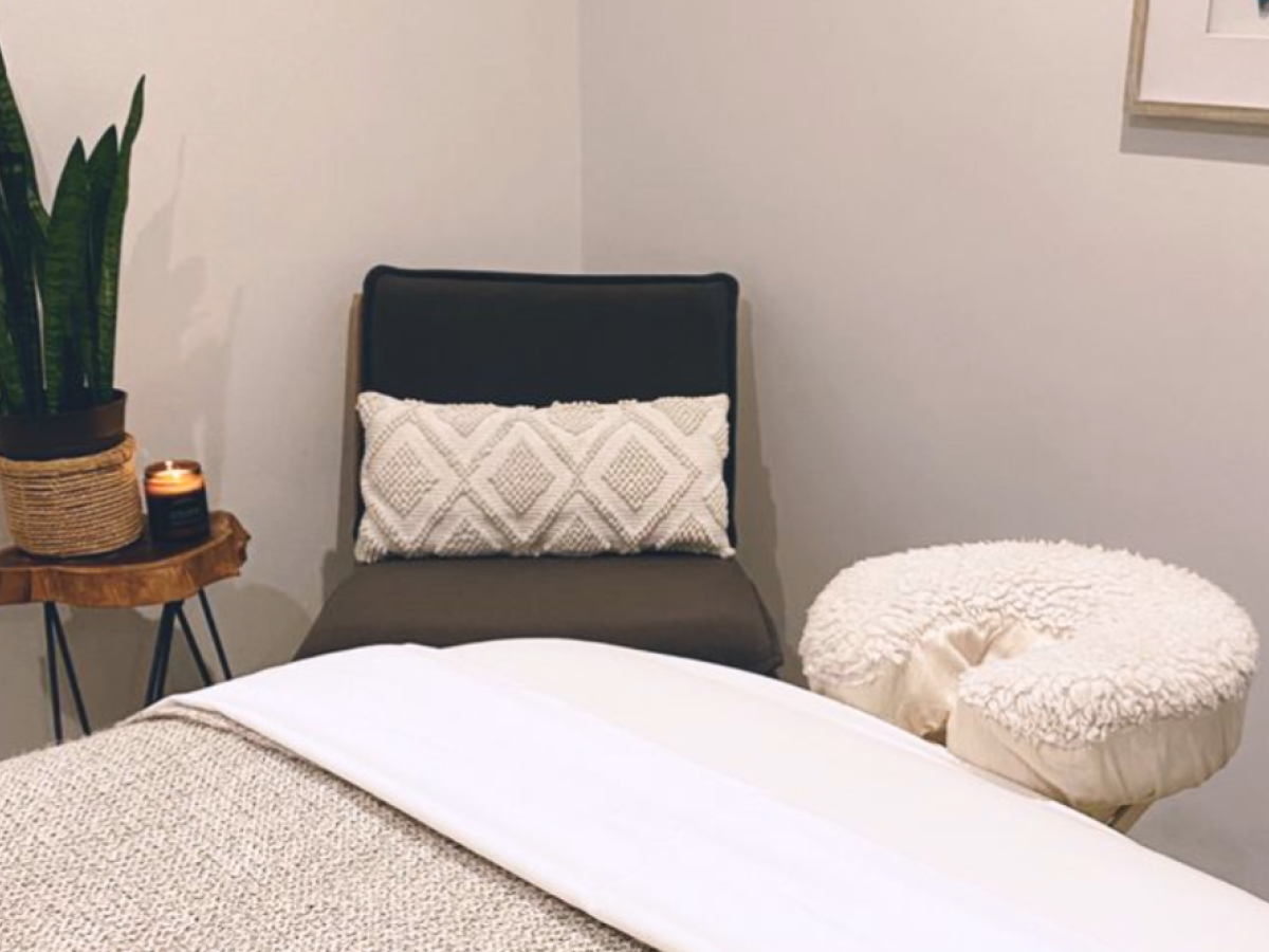A cozy room with a chair, cushion, small table, candle, plant, and massage table with white sheets, creating a peaceful ambiance.