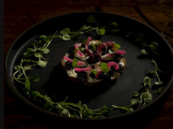 A dark plate with a decorative dish featuring assorted colorful ingredients, garnished with greens, artfully presented for a gourmet experience.