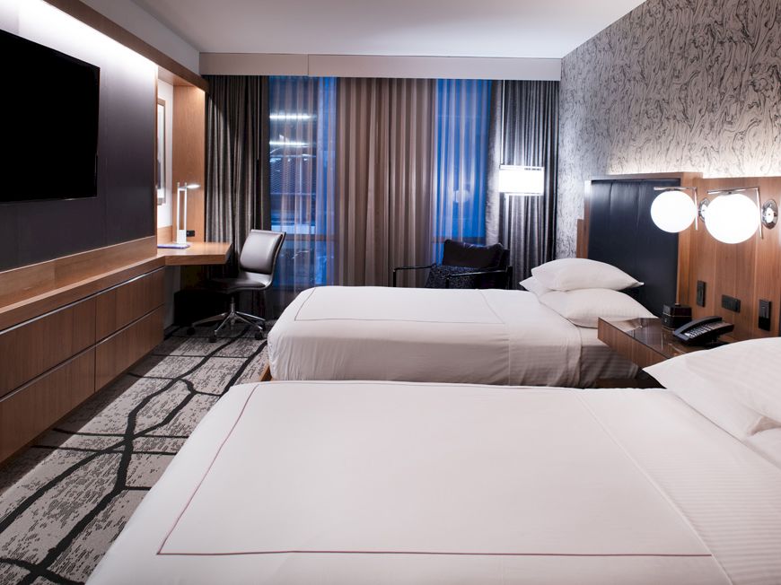 The image shows a hotel room with two beds, modern decor, a desk, a chair, a TV on the wall, and stylish lighting.