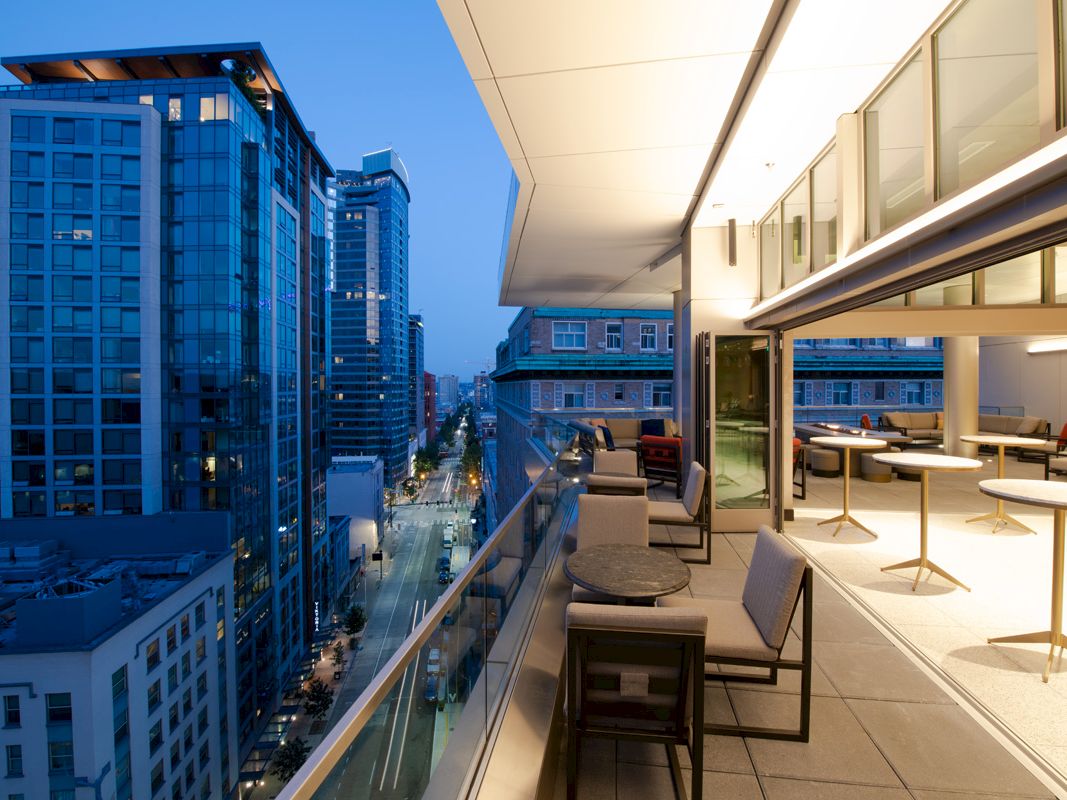 A modern rooftop terrace overlooking a cityscape at dusk, featuring seating and tables, with tall buildings visible in the background.