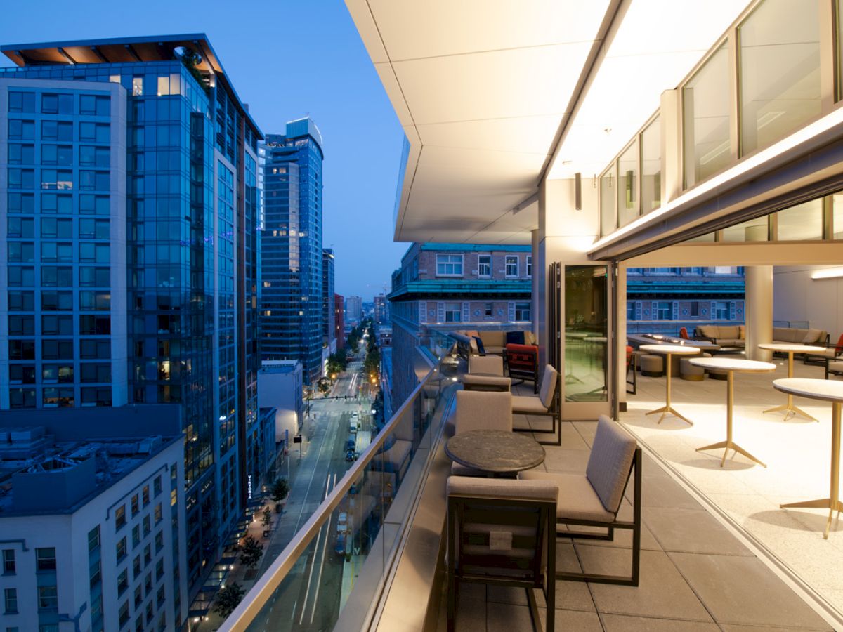 A modern outdoor terrace overlooking a cityscape at dusk, with seating and tables arranged alongside a glass railing.