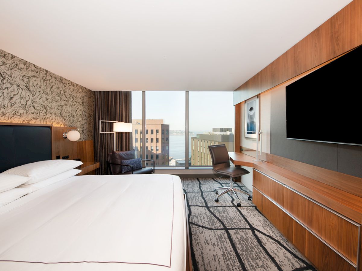 A modern hotel room with a large bed, desk, and chair, featuring a wall-mounted TV and a city view through wide windows.