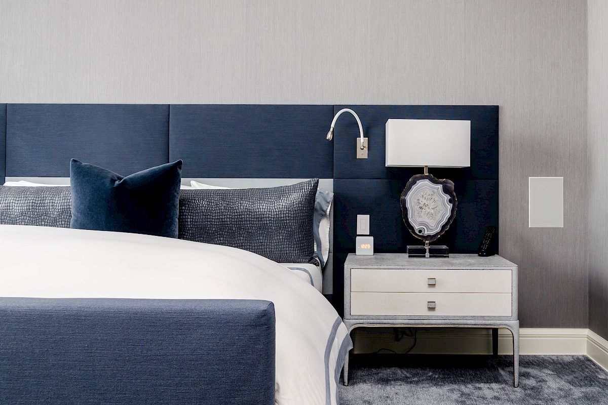 A modern bedroom with a navy bed, white bedding, gray carpet, nightstand with a lamp, clock, and built-in wall light is shown here.