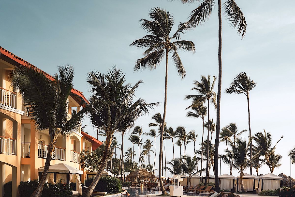 A beautiful resort pool with lounge chairs, surrounded by tall palm trees and a multi-story building under a clear sky.